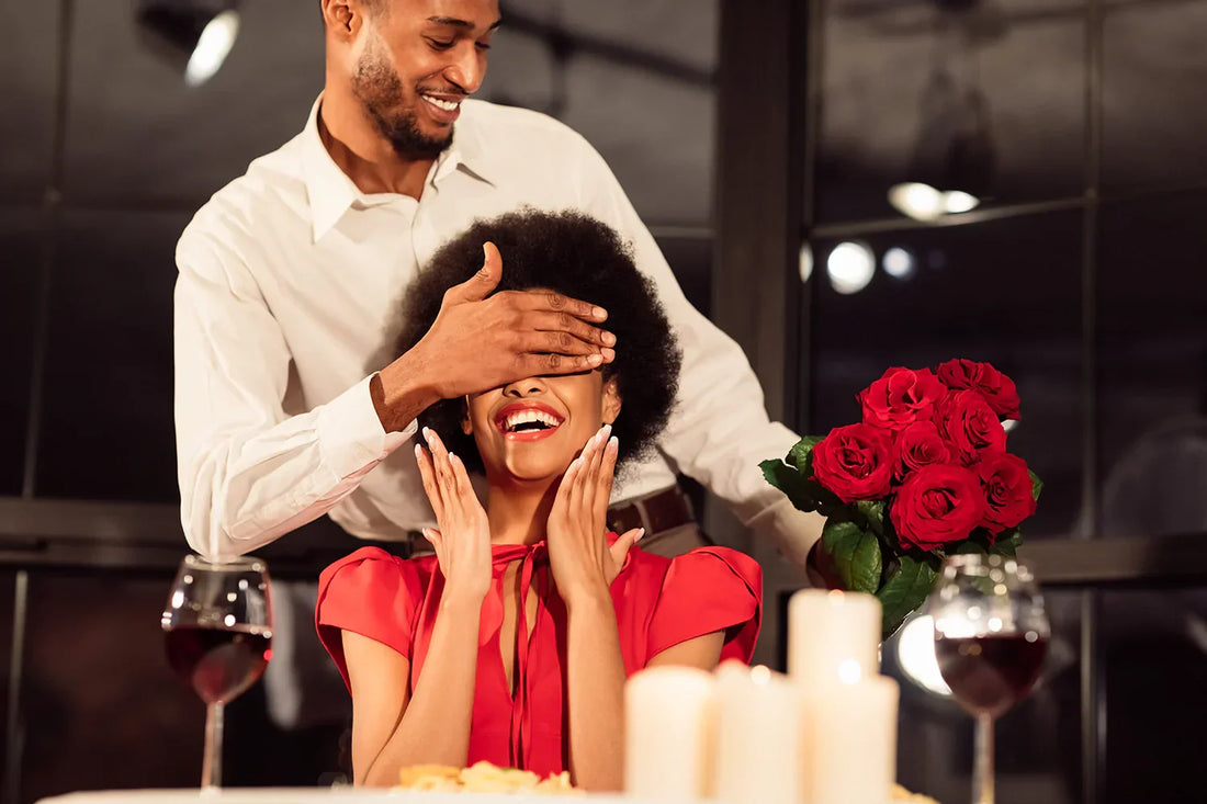 Smiling man surprises his partner with bouquet of long-stemmed red roses at a candle-lit dinner